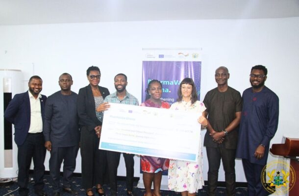 Group of eight people at an award event holding a large ceremonial check, standing in a room with a banner in the background and a crest in the corner.