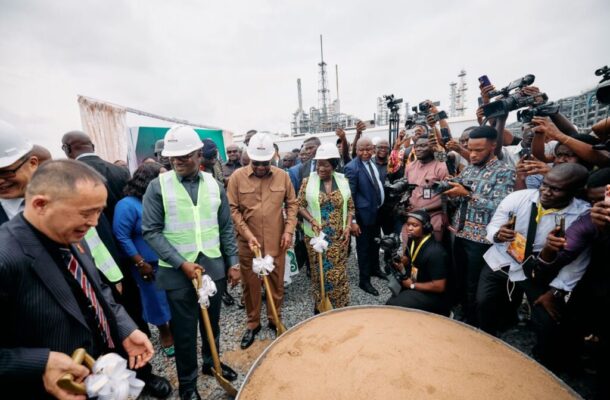 Officials in hard hats and safety vests breaking ground with ceremonial shovels while photographers crowd around at a construction site groundbreaking ceremony.