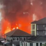 Large fire in an urban area with bright orange flames and dark smoke rising behind low-rise buildings.