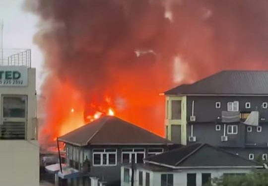 Large fire in an urban area with bright orange flames and dark smoke rising behind low-rise buildings.
