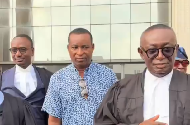 Three men stand outside a glass-fronted building; two wear dark legal robes with white collars, one in a blue patterned shirt and sunglasses hanging from his chest.