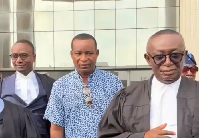 Three men stand outside a glass-fronted building; two wear dark legal robes with white collars, one in a blue patterned shirt and sunglasses hanging from his chest.