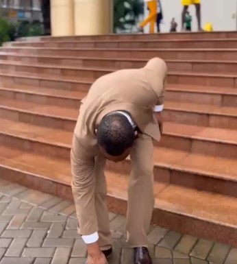 Man in a tan suit bends over on outdoor wooden stairs, touching his shoe.