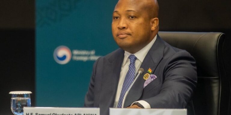 Man in a dark suit and blue patterned tie sits at a panel table with a microphone, water glass, and a nameplate in front, during a formal conference.