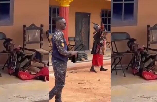 Uniformed security officer stands with a large object while a person in red pants gestures nearby; someone lies on the ground outside a yellow building with chairs nearby.
