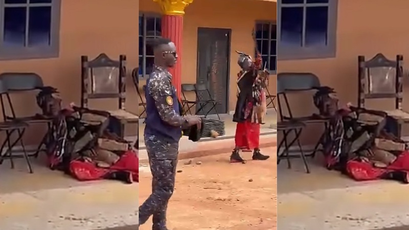 Uniformed security officer stands with a large object while a person in red pants gestures nearby; someone lies on the ground outside a yellow building with chairs nearby.