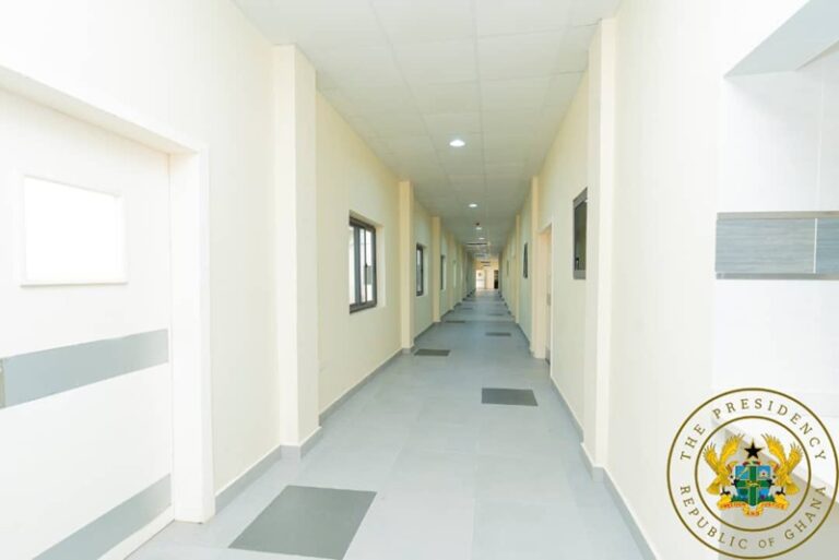 Long, well-lit corridor in a modern building with doors and windows along both sides; crest of the Ghana Presidency visible in the bottom-right corner.