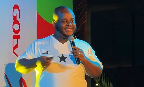 Man on stage speaking into a handheld microphone, wearing a white shirt with a black star emblem and a Puma logo visible on the chest.