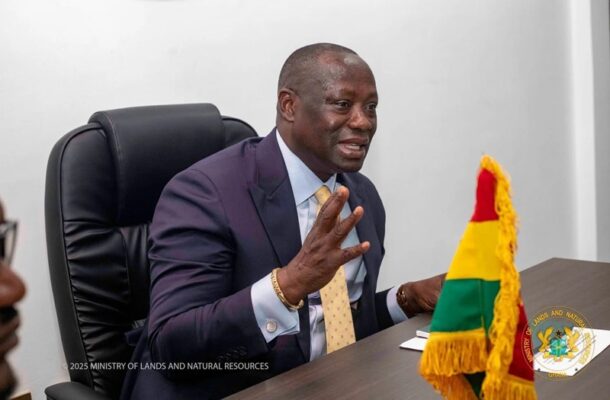 A man in a navy suit and yellow tie speaks at a desk, gesturing with his right hand in a meeting, with a Ghana flag on the table and an official seal nearby.