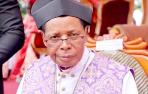 Religious cleric in ornate purple vestments and a square hat, wearing glasses at an outdoor ceremony.