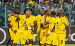 A group of soccer players in bright yellow uniforms celebrate on the field as teammates high-five and gesture, with fans in the stands behind them.