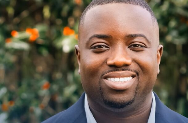 Portrait of a smiling Black man in a navy blazer and white shirt outdoors, with greenery in the background.