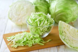 Shredded green cabbage in a glass bowl with chopped cabbage on a wooden cutting board and whole heads in the background