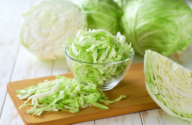 Shredded green cabbage in a glass bowl with chopped cabbage on a wooden cutting board and whole heads in the background
