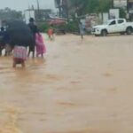 People wading through knee-deep floodwaters on a city street, some under umbrellas, with cars halted in the background.