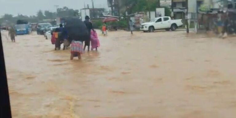 People wading through knee-deep floodwaters on a city street, some under umbrellas, with cars halted in the background.