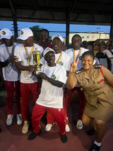 A group of men in white shirts and red pants with medals, posing with a trophy; a smiling woman in uniform shows a peace sign beside them.