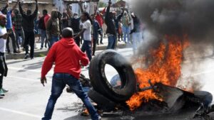Crowd of protesters on a street raising arms as a large burning tire and debris blaze on the road with smoke rising from the fire.