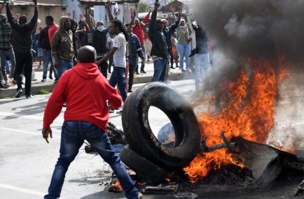 Crowd of protesters on a street raising arms as a large burning tire and debris blaze on the road with smoke rising from the fire.