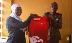 Smiling woman in a white hijab hands a red sports jersey to a uniformed man indoors against a yellow wall.