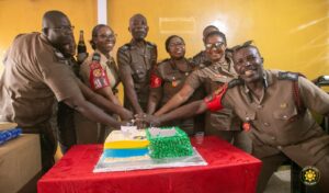 Group of uniformed police officers with hands joined over a decorated cake at a celebration, indoors against a yellow wall.
