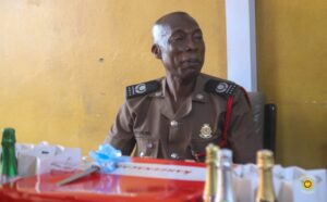 Senior police officer in a tan uniform sits at a table with gifts and bottles on a yellow wall backdrop.