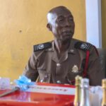 Uniformed police officer seated at a table with wrapped gifts and bottles, against a yellow wall.