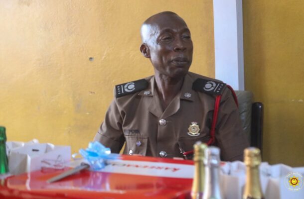Uniformed police officer seated at a table with wrapped gifts and bottles, against a yellow wall.