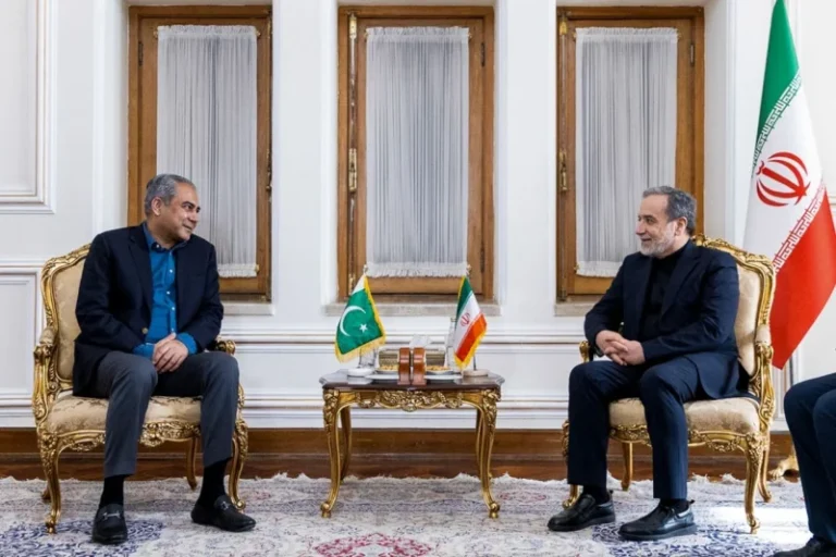 Two men in suits sit opposite each other in an ornate room; a small table between them holds Pakistan and Iran flags.