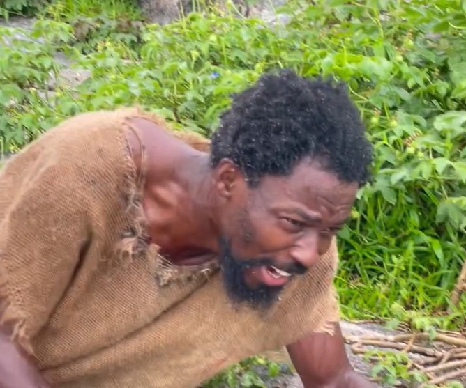 Man with a burlap tunic leaning forward outdoors, surrounded by green bushes, appearing tired or distressed.