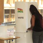 Voter casts ballot at a white polling booth labeled VOTE, with a Ghana flag sticker nearby.