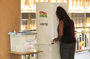 Voter casts ballot at a white polling booth labeled VOTE, with a Ghana flag sticker nearby.