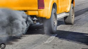 Rear of a yellow taxi emitting dark exhaust smoke as it drives on a road.