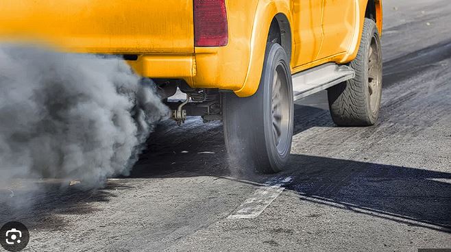 Rear of a yellow taxi emitting dark exhaust smoke as it drives on a road.
