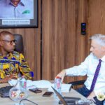 Two men in a formal office meeting, seated at a large desk with documents, water bottles, and a laptop; a Ghana flag and framed portrait hang on a wood-paneled wall behind them.