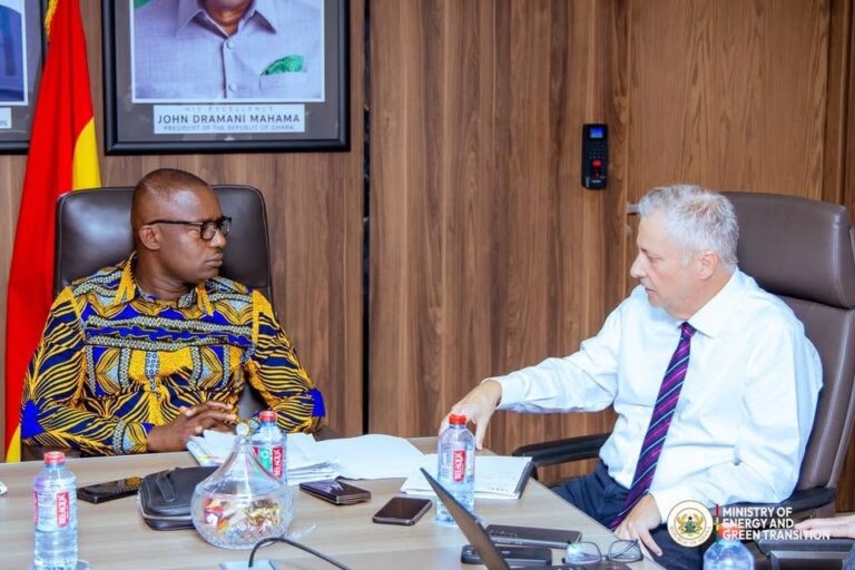 Two men in a formal office meeting, seated at a large desk with documents, water bottles, and a laptop; a Ghana flag and framed portrait hang on a wood-paneled wall behind them.