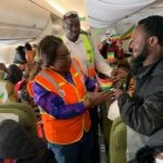Passengers in an airplane aisle, including a woman in an orange safety vest, hand a small colorful flag to a smiling man in a black jacket as others watch and clap in the background.