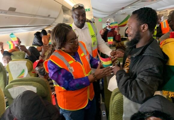 evacuees | Ghanaguardian.com Passengers in an airplane aisle, including a woman in an orange safety vest, hand a small colorful flag to a smiling man in a black jacket as others watch and clap in the background.