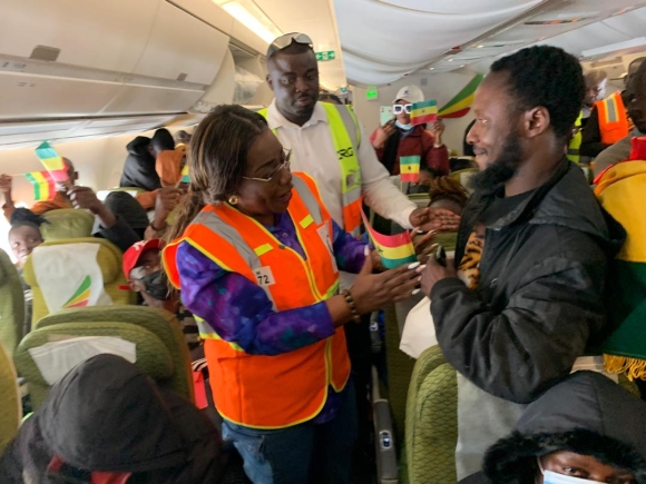 Passengers in an airplane aisle, including a woman in an orange safety vest, hand a small colorful flag to a smiling man in a black jacket as others watch and clap in the background.