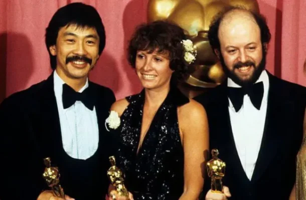 Three casually smiling individuals in formal attire pose with Oscar statues at a red-carpet event, each holding a trophy.