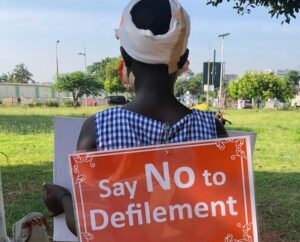 Person with a bandaged head facing away holds an orange poster that says 'Say No to Defilement' in a sunny park area.