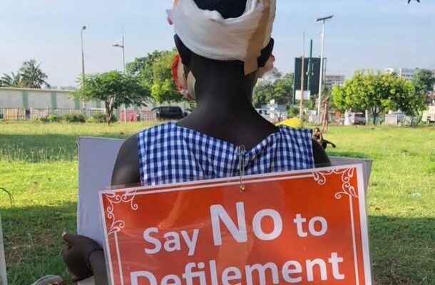 file photo | Ghanaguardian.com Person with a bandaged head facing away holds an orange poster that says 'Say No to Defilement' in a sunny park area.