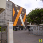 Exterior view of the Ministry of Finance building with orange-brown geometric panels and a crest, gated entrance, and surrounding trees in Ghana