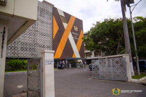 Exterior view of the Ministry of Finance building with orange-brown geometric panels and a crest, gated entrance, and surrounding trees in Ghana