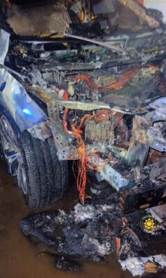 Front end of a severely burned car with melted wiring and charred debris exposed at the engine bay (fire damage).