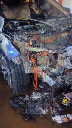 Front end of a severely burned car with melted wiring and charred debris exposed at the engine bay (fire damage).