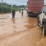 Flooded road scene with muddy water; a white SUV is partially submerged on the right as two people stand in the water ahead near a red truck.