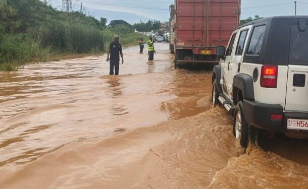 Flooded road scene with muddy water; a white SUV is partially submerged on the right as two people stand in the water ahead near a red truck.
