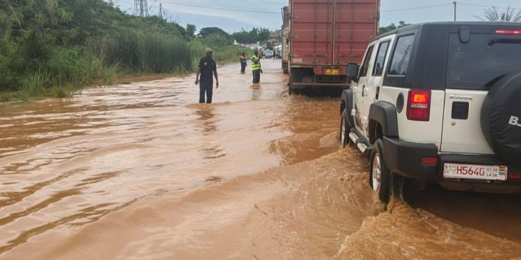 Flooded road scene with muddy water; a white SUV is partially submerged on the right as two people stand in the water ahead near a red truck.