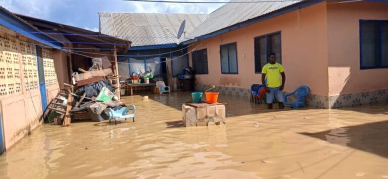 Flooded courtyard of an orange house with muddy water covering the ground; a man in a yellow shirt stands near blue chairs while buckets sit on a table.